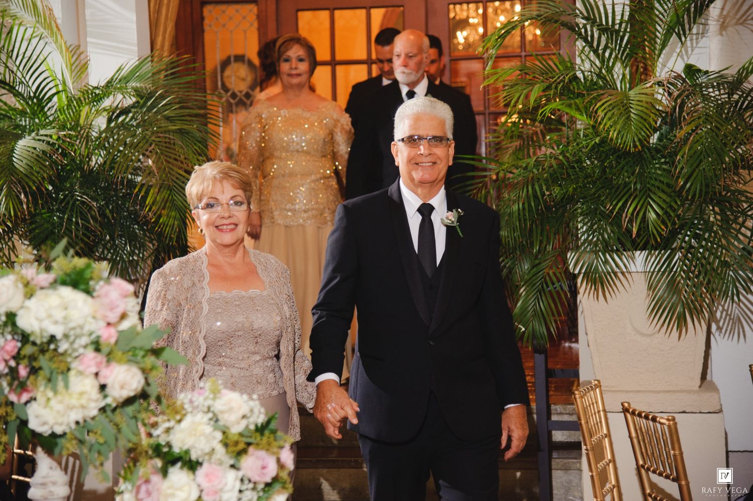 Elegant couple walking hand-in-hand down the aisle at a wedding ceremony surrounded by tropical palms.