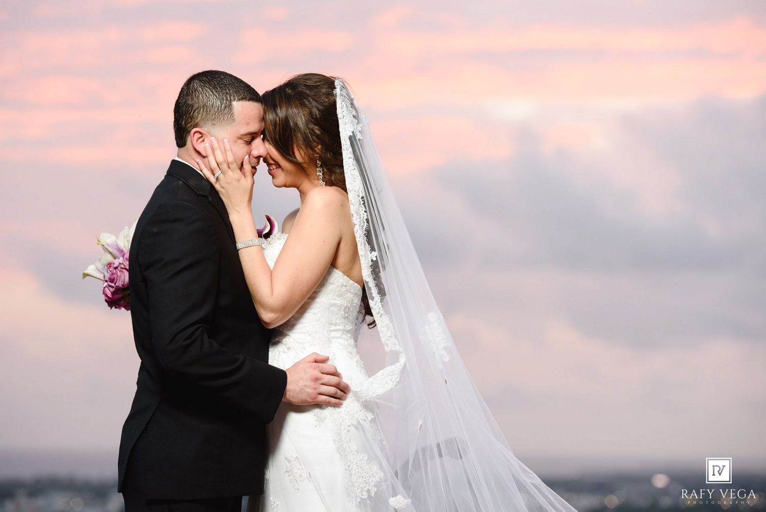 Bride and groom sharing a romantic kiss at sunset with a pink and purple sky backdrop on their wedding day.