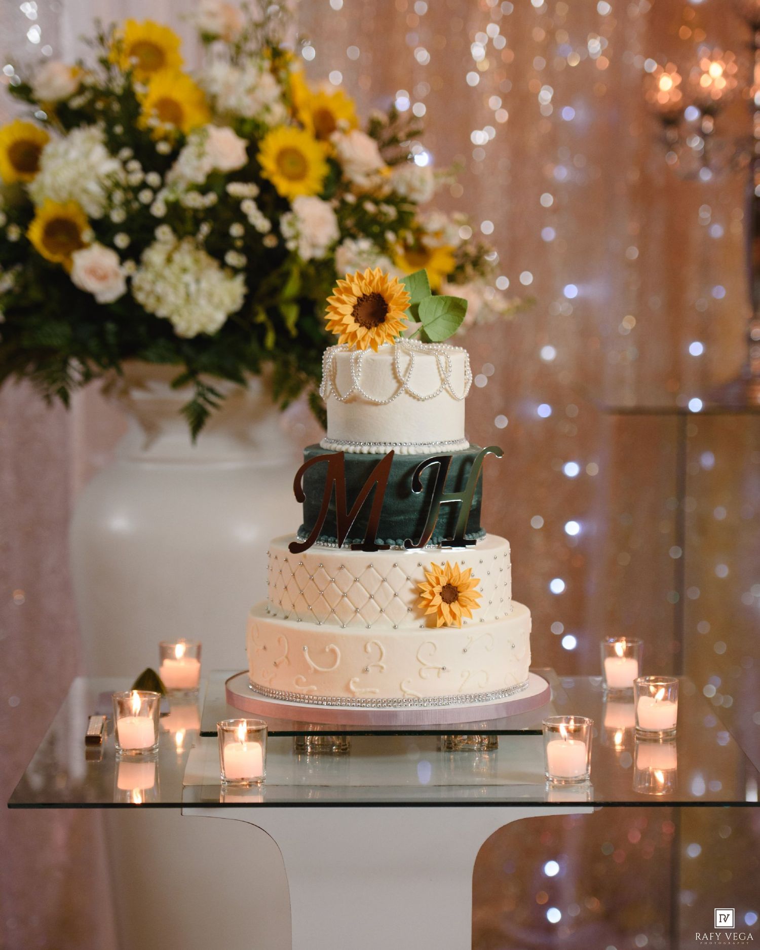 Three-tier white wedding cake with sunflower accents and monogram, surrounded by candles and floral arrangements.