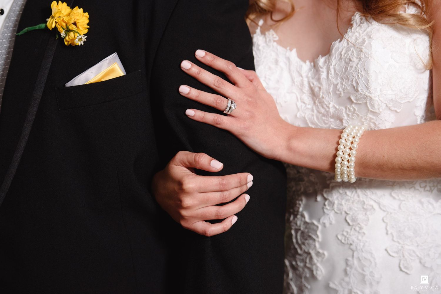 Bride and groom closeup showing wedding rings, pearl bracelet, and yellow floral boutonniere on black tuxedo.