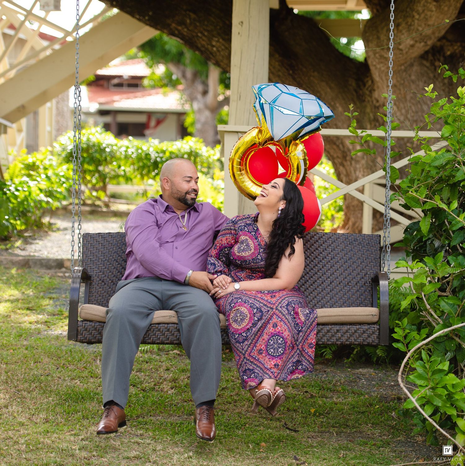 Couple sitting on a swing holding hands with a diamond ring balloon, celebrating their engagement outdoors.
