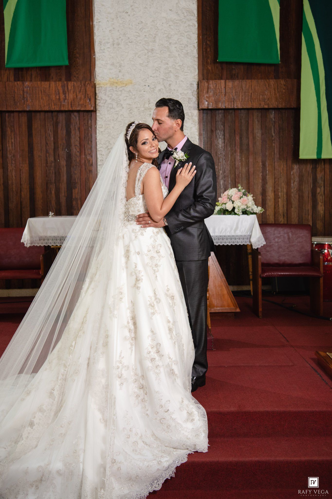 Bride and groom share a kiss at the altar of a wooden church, bride wearing a ballgown with long veil.