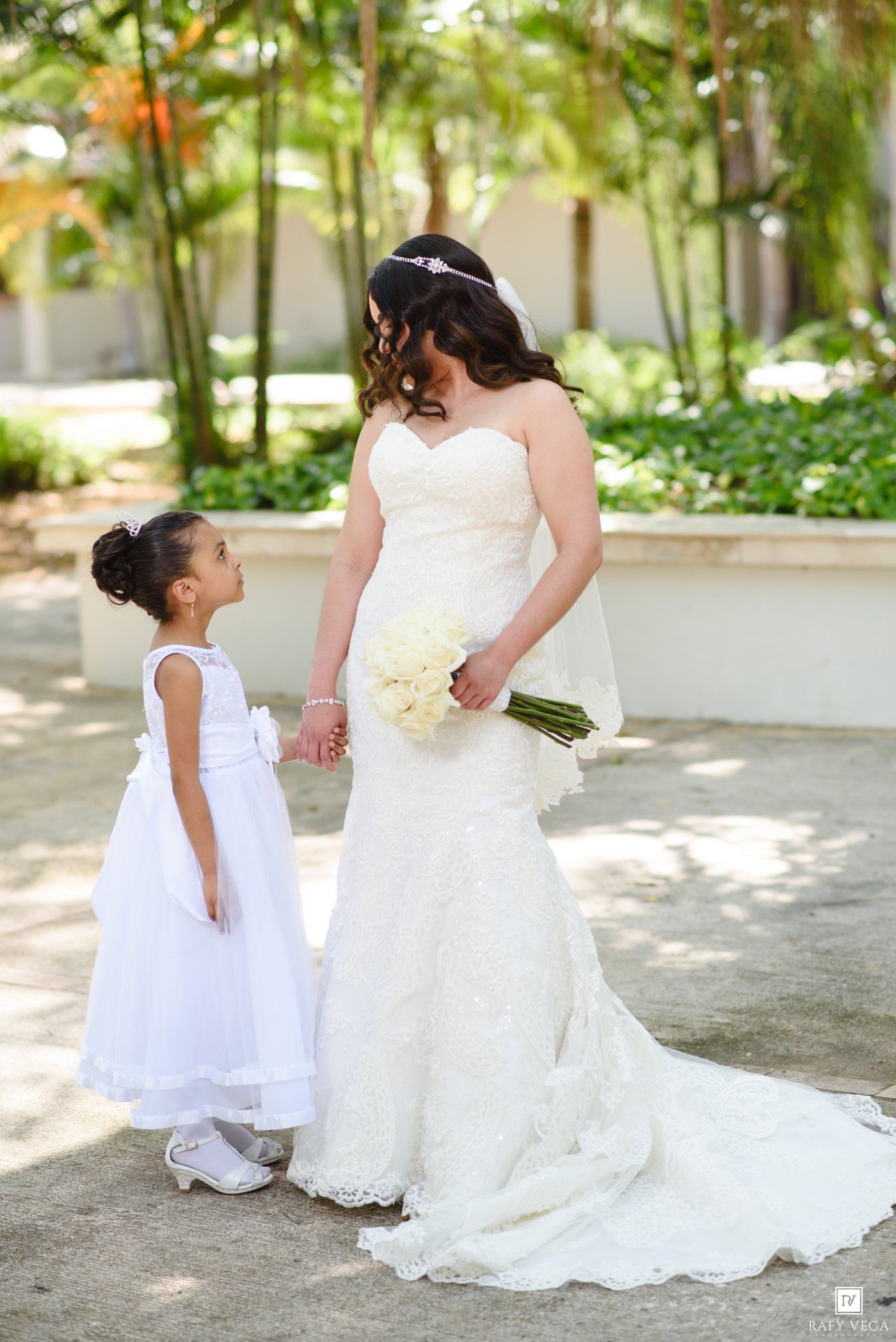 Bride in lace wedding gown holds white bouquet while flower girl in white dress looks up at her outdoors.
