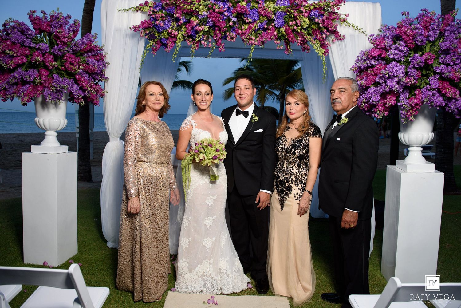 Wedding party of five posing under a floral arch with purple flowers at an outdoor beachside ceremony.