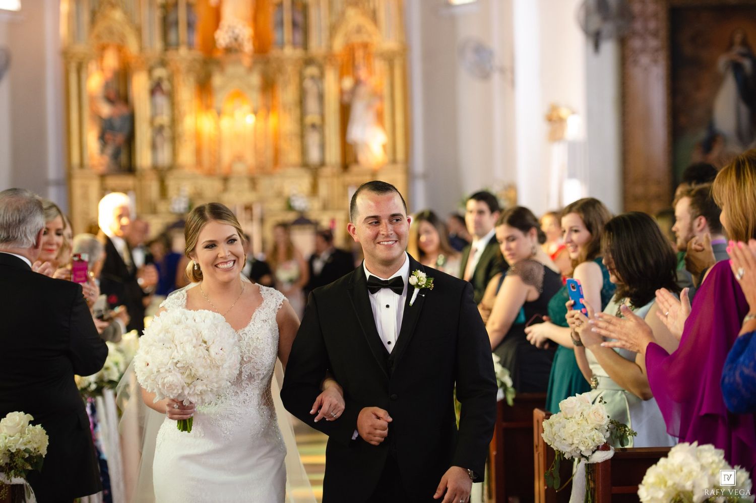 Bride and groom walk down the aisle of an ornate church, smiling as guests applaud their wedding ceremony.