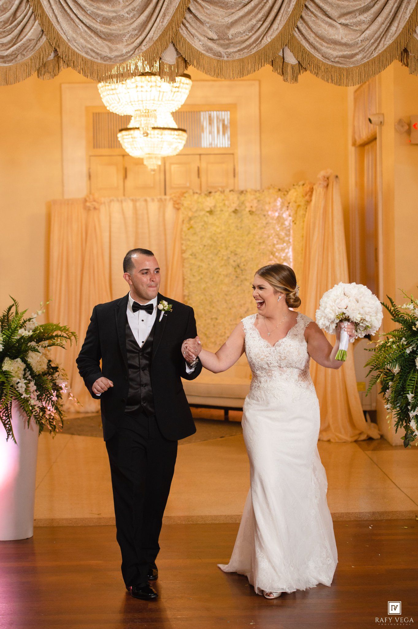 Joyful newlyweds walk hand-in-hand at elegant wedding reception, bride in lace gown holding white bouquet, groom in black tuxedo.