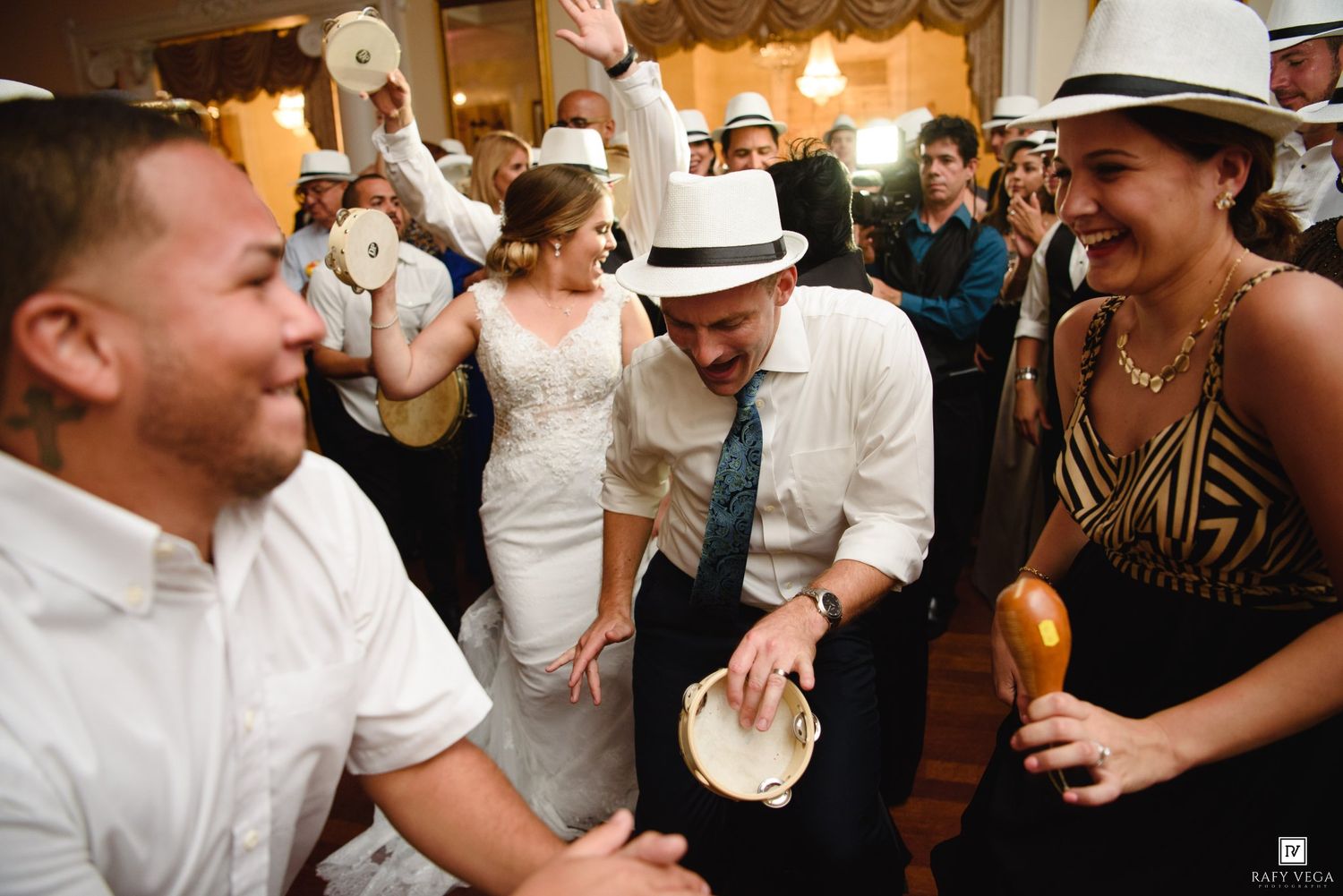 Wedding guests dancing joyfully at a reception, with a groom in a white hat playing maracas while a bride watches in the background.