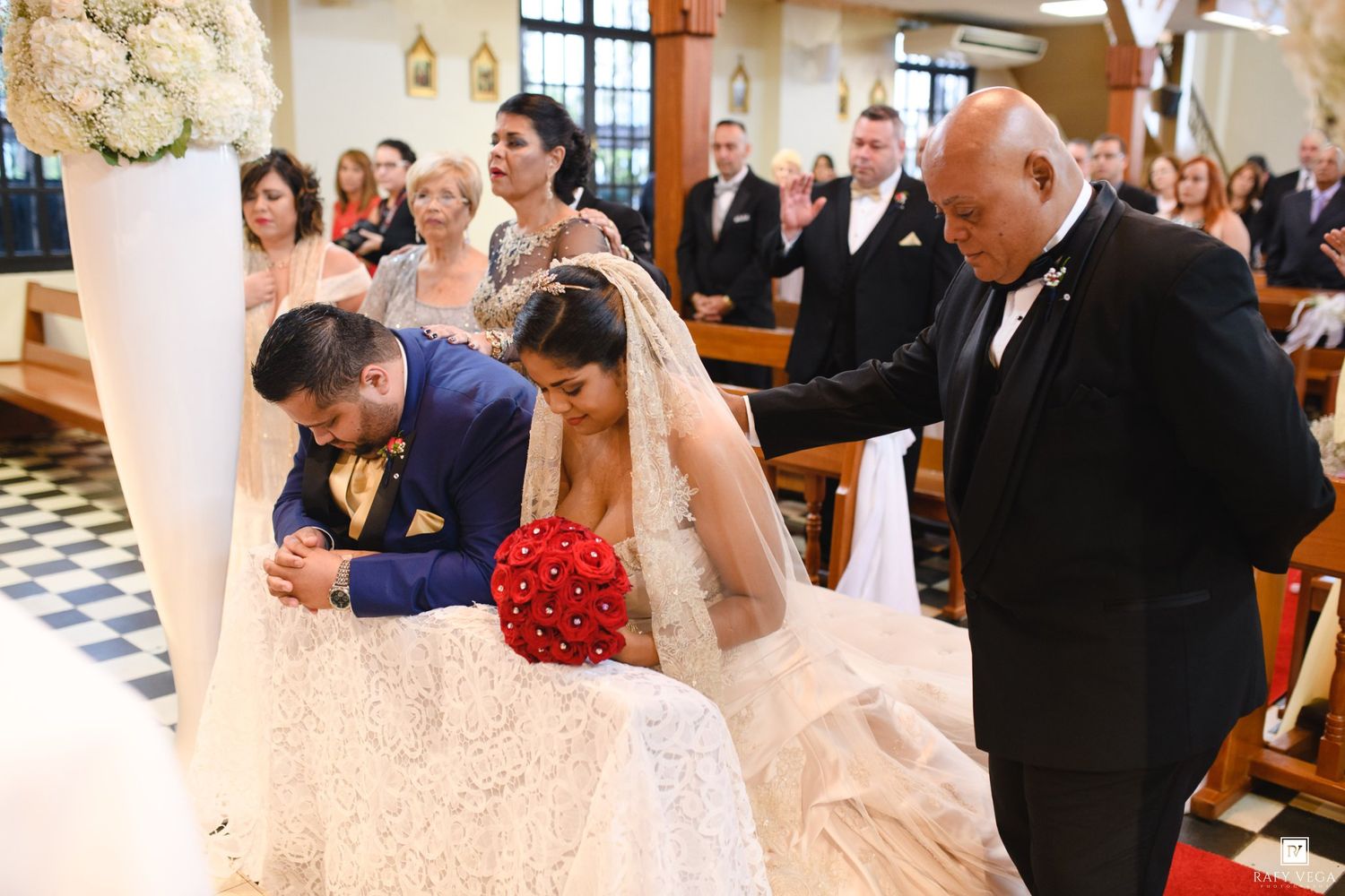 Bride and groom kneeling at church altar during wedding ceremony with guests and officiant in background.