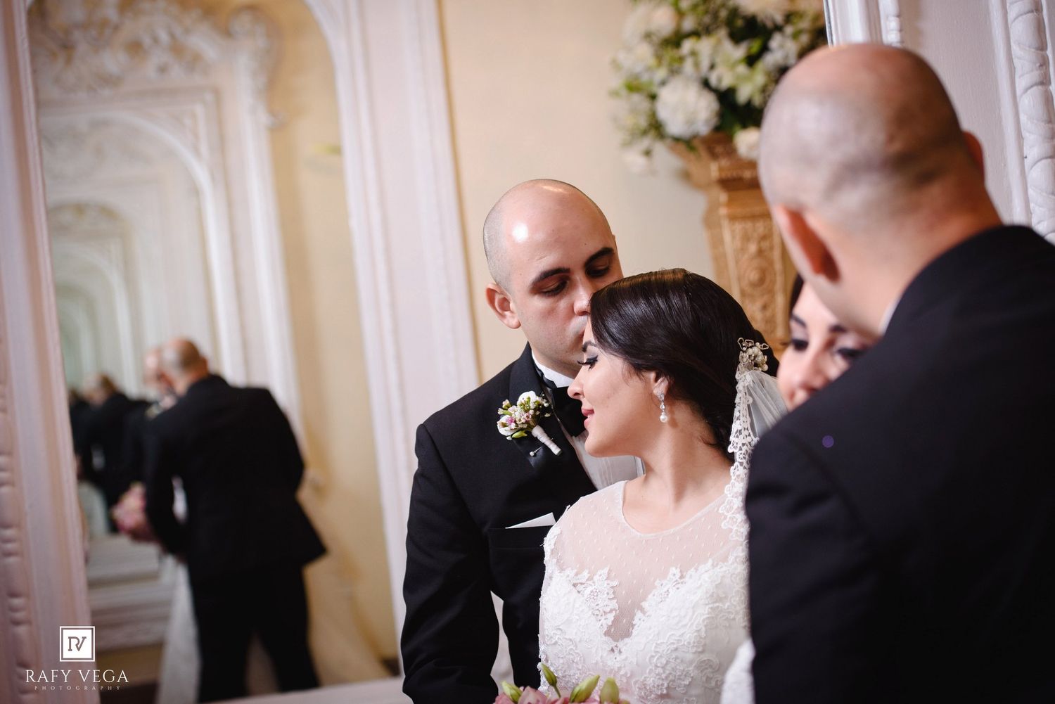 A groom kisses his bride on the forehead during their elegant wedding ceremony in a grand ornate venue.