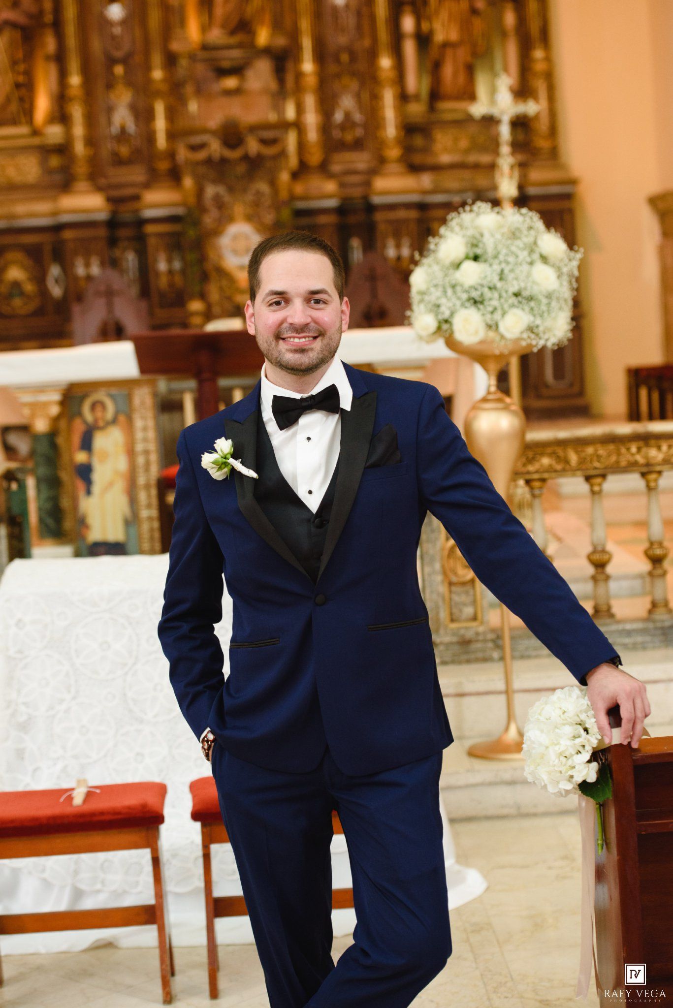 Smiling groom in navy blue tuxedo with boutonniere posing in elegant church before wedding ceremony.