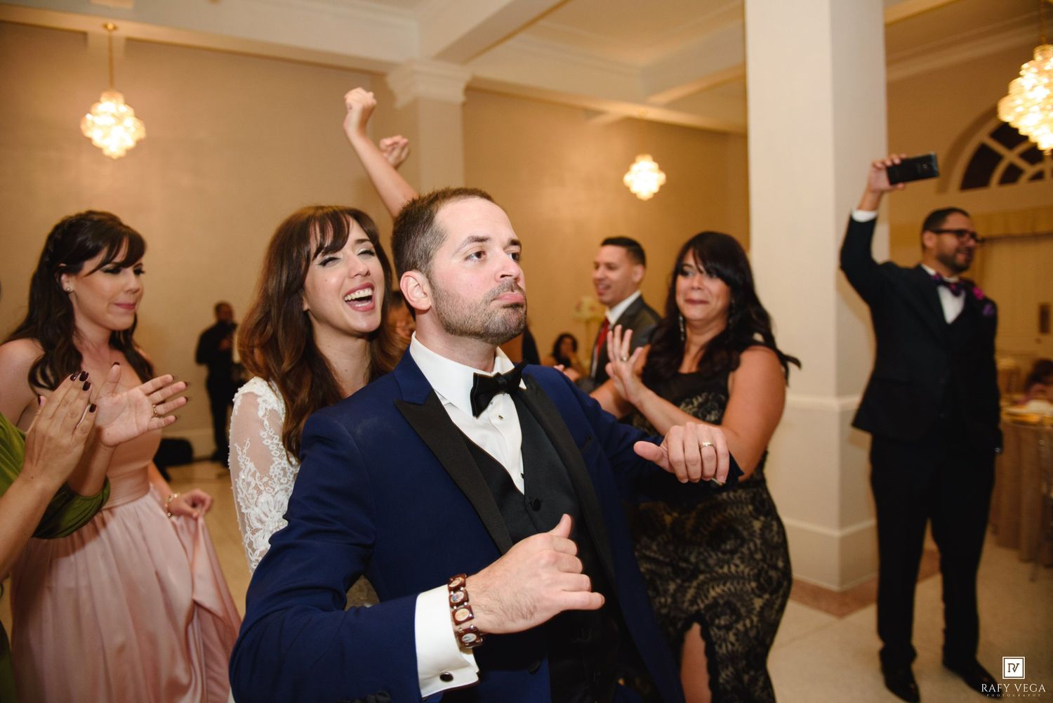 Guests celebrate joyfully at a wedding reception, clapping and dancing in an elegant venue.