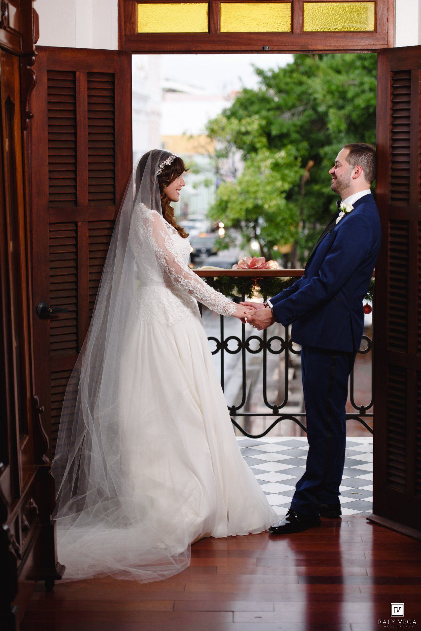 Bride in white gown and veil holds hands with groom in navy suit on elegant balcony doorway during wedding ceremony.