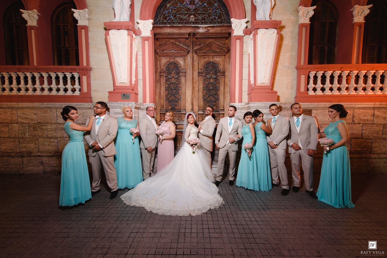 Wedding party poses outside a church, bride in white gown flanked by groomsmen in tan suits and bridesmaids in teal dresses.