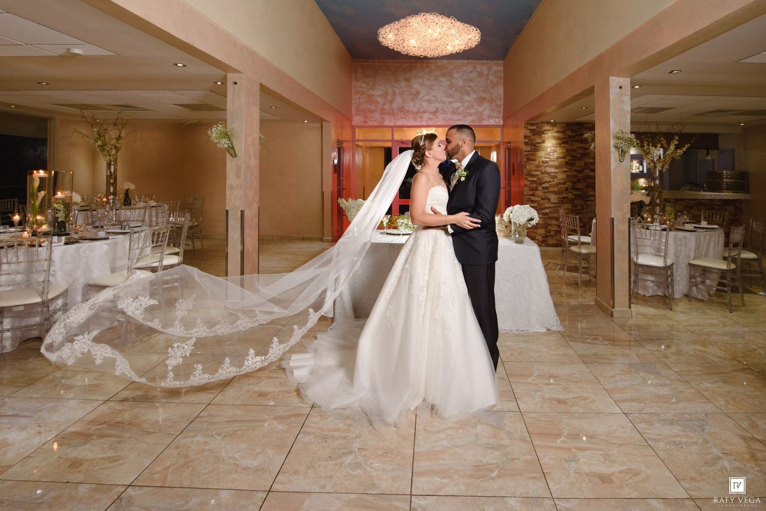 Bride and groom share a kiss in a beautifully decorated wedding venue with a flowing lace veil.
