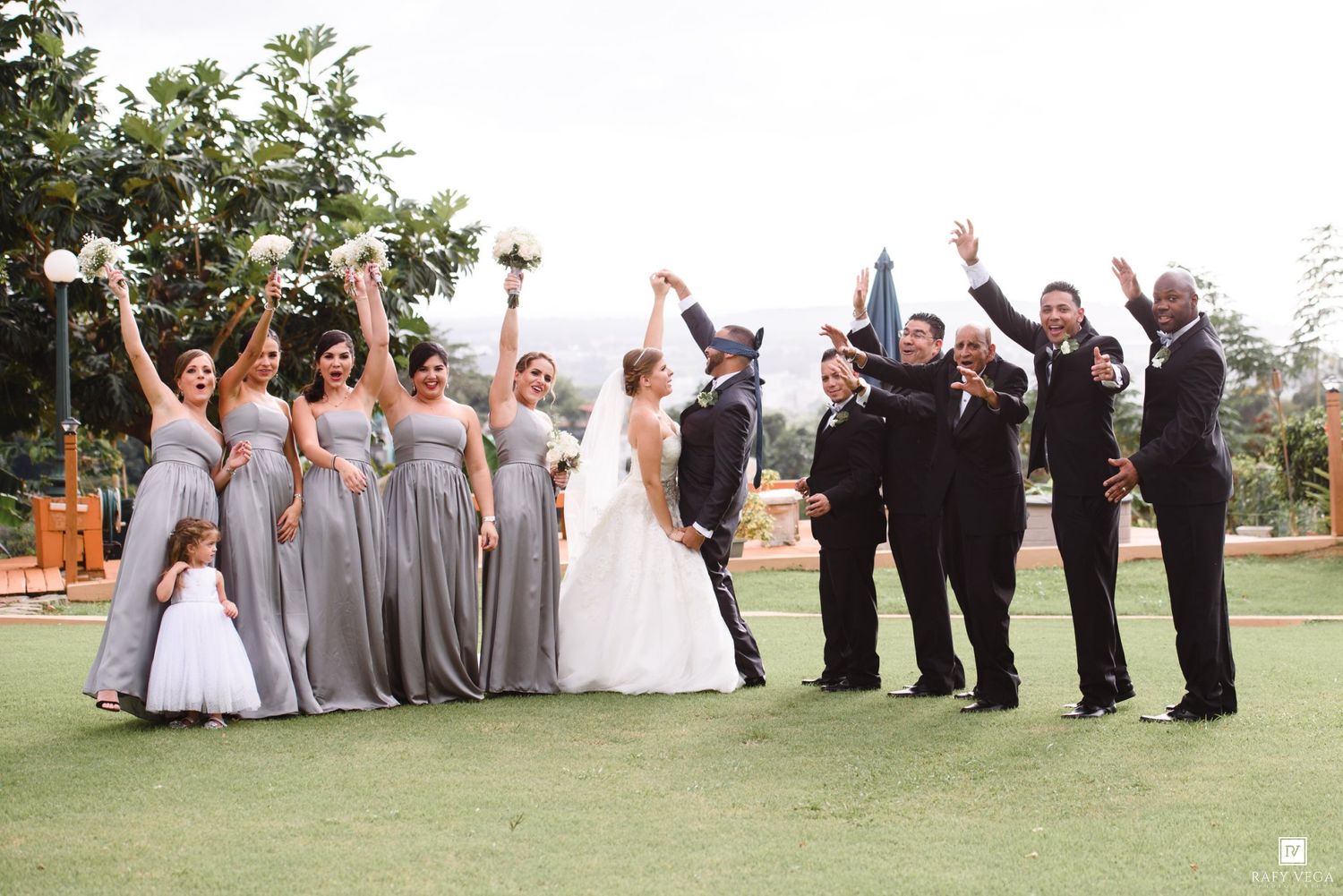 Joyful wedding party celebrating with raised arms outdoors, bridesmaids in gray gowns and groomsmen in black tuxedos.