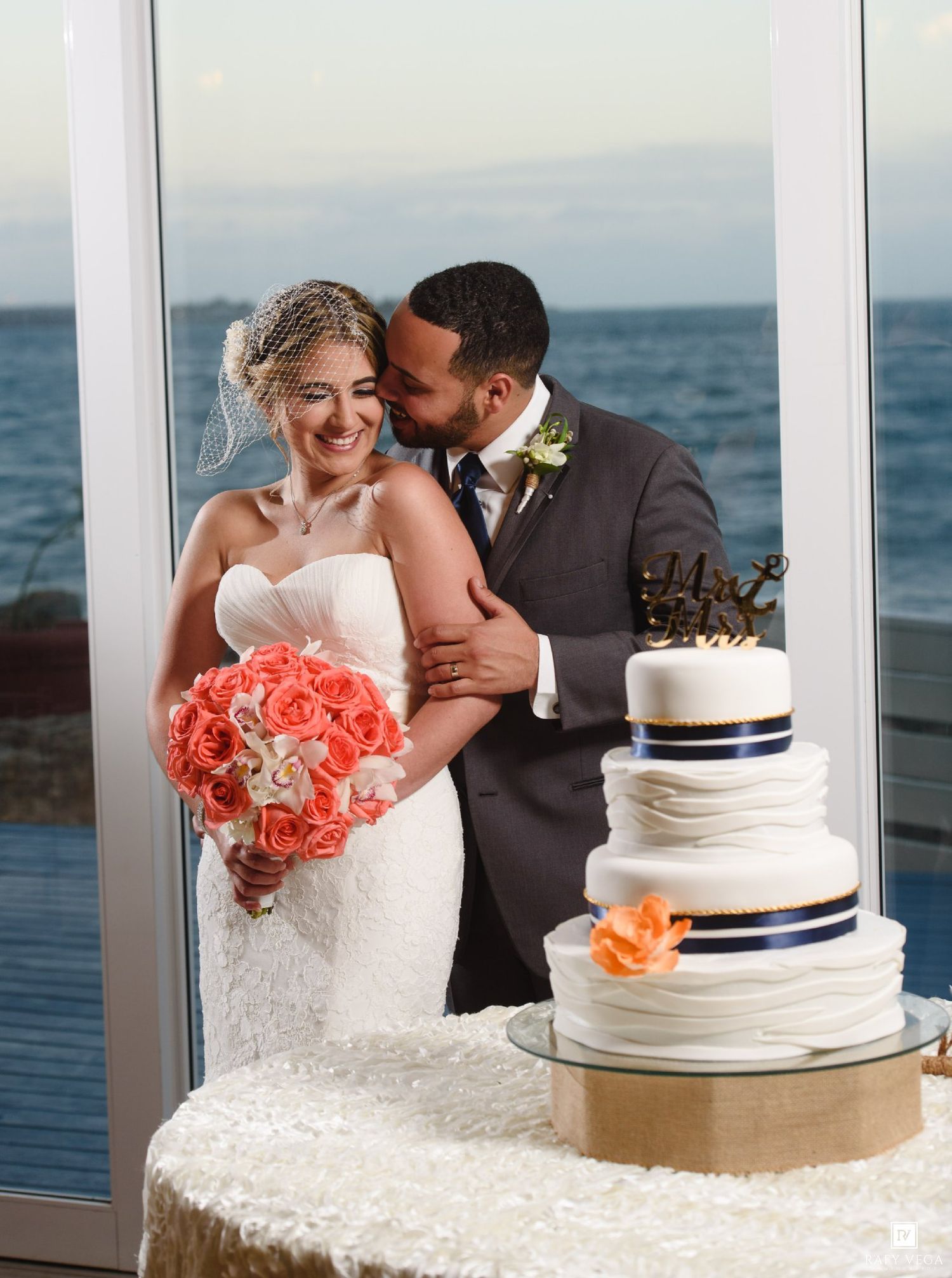 Bride and groom smiling beside three-tier wedding cake with ocean view backdrop.