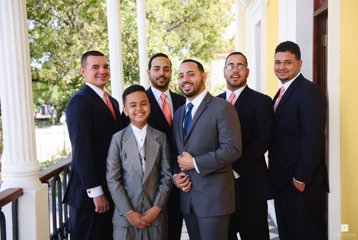 Group of six men in formal suits posing together on a porch with white columns at a wedding celebration.