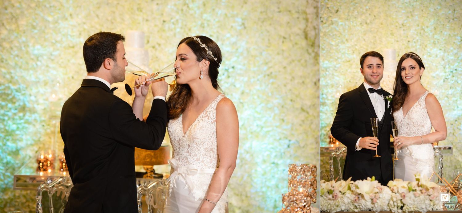 Bride and groom share a champagne toast at their elegant wedding reception with a floral backdrop.