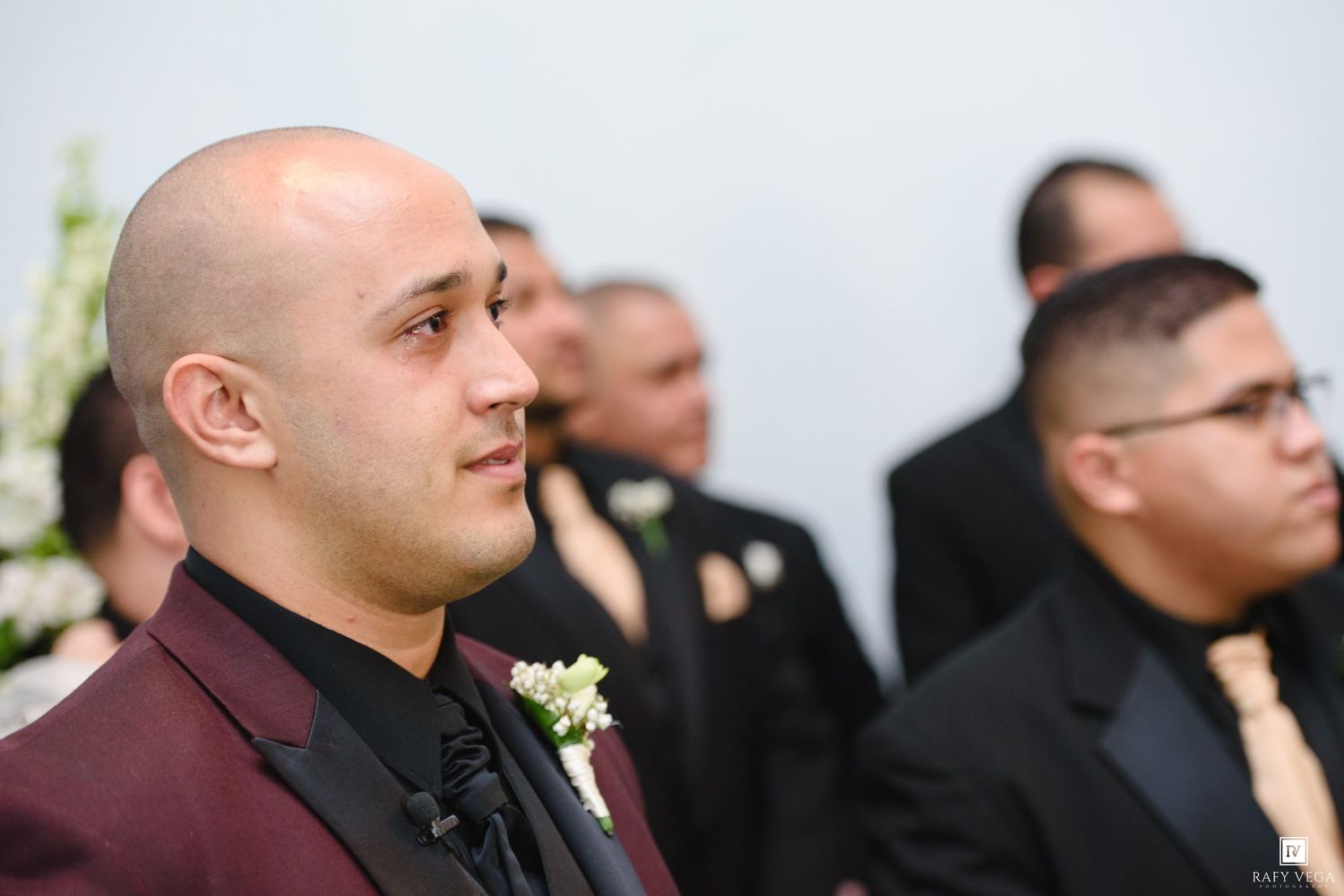 Groom in burgundy suit with boutonniere stands at wedding altar, groomsmen in black suits visible behind him.