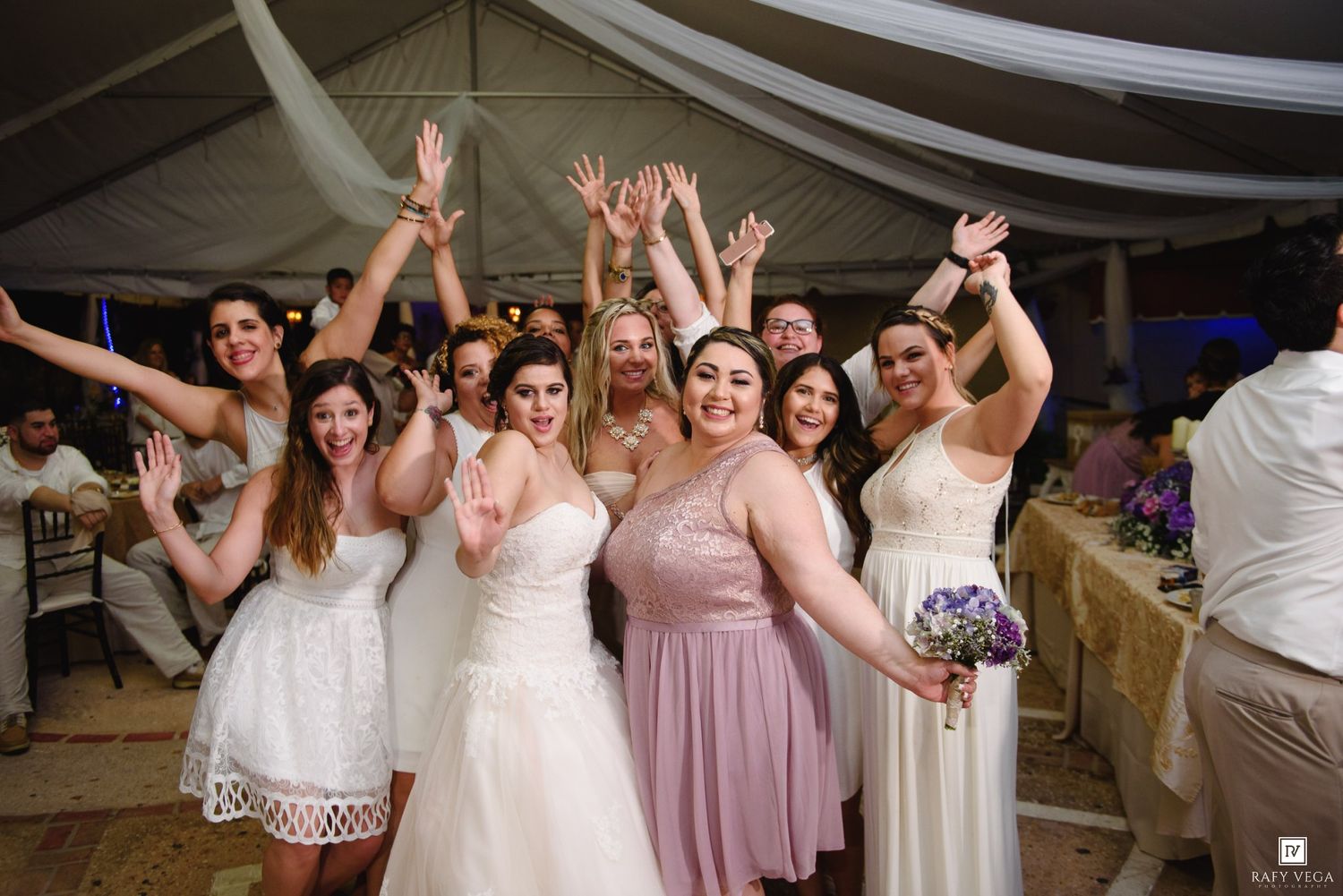 Bride and bridesmaids celebrating joyfully at wedding reception under tent, raising hands and smiling.