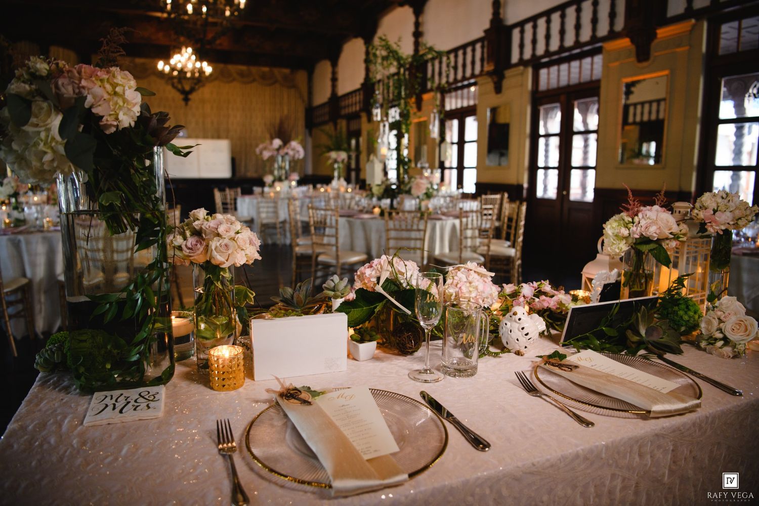 Elegant wedding reception table with pink hydrangeas, greenery centerpieces, gold accents, and white linens in a rustic ballroom.