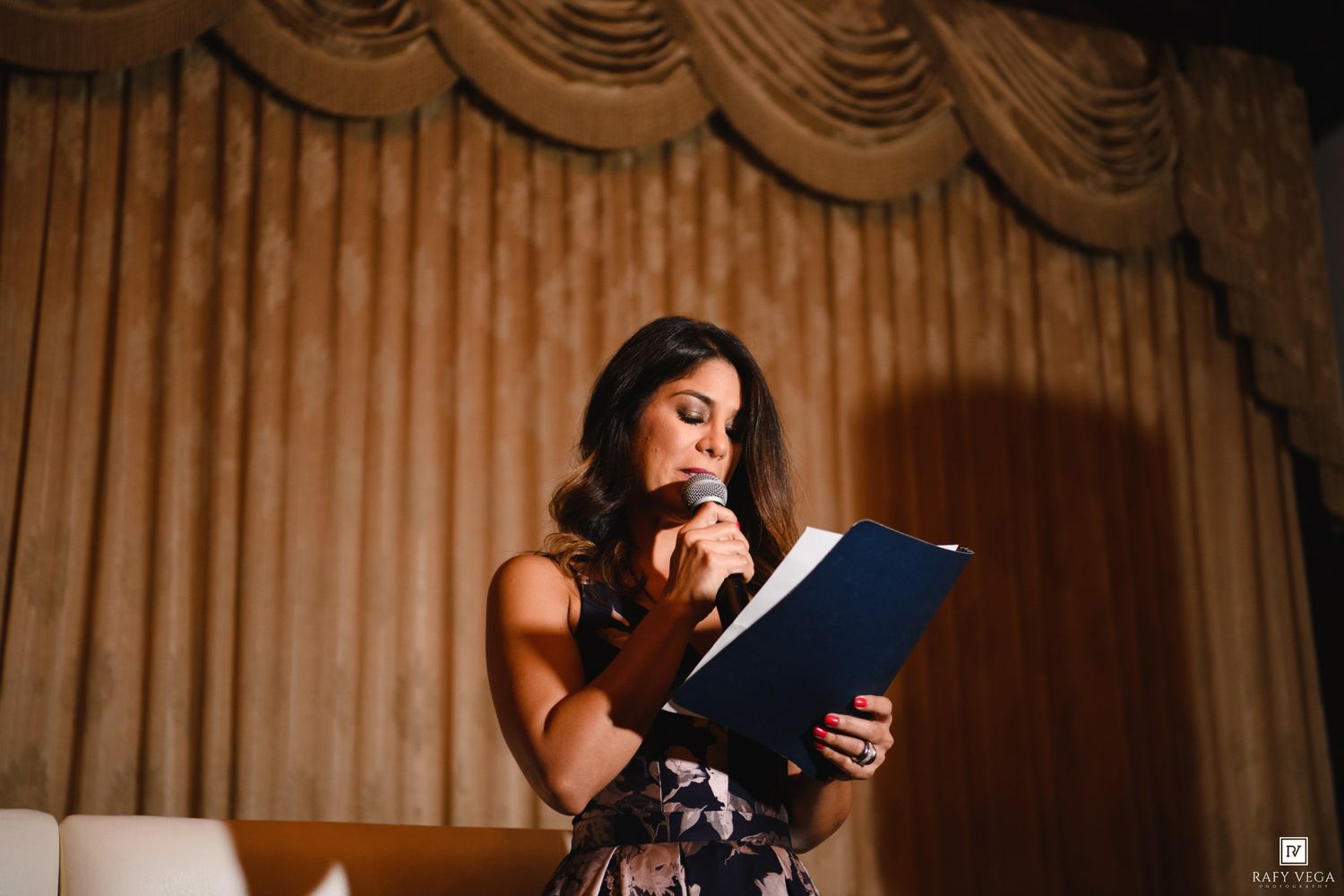 A woman in a floral dress reads from a blue folder while speaking into a microphone on stage.