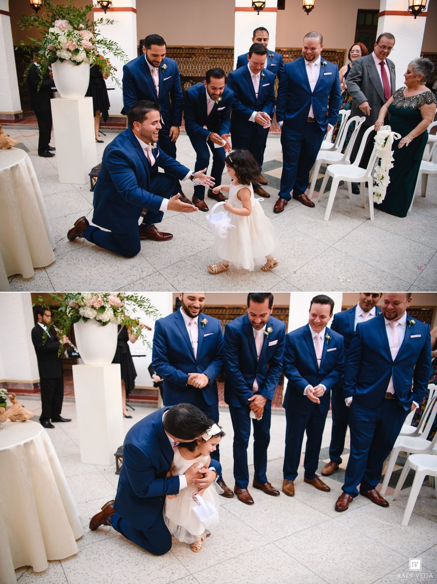 Groomsmen in navy suits kneel to interact with adorable flower girl in white dress during outdoor wedding ceremony.