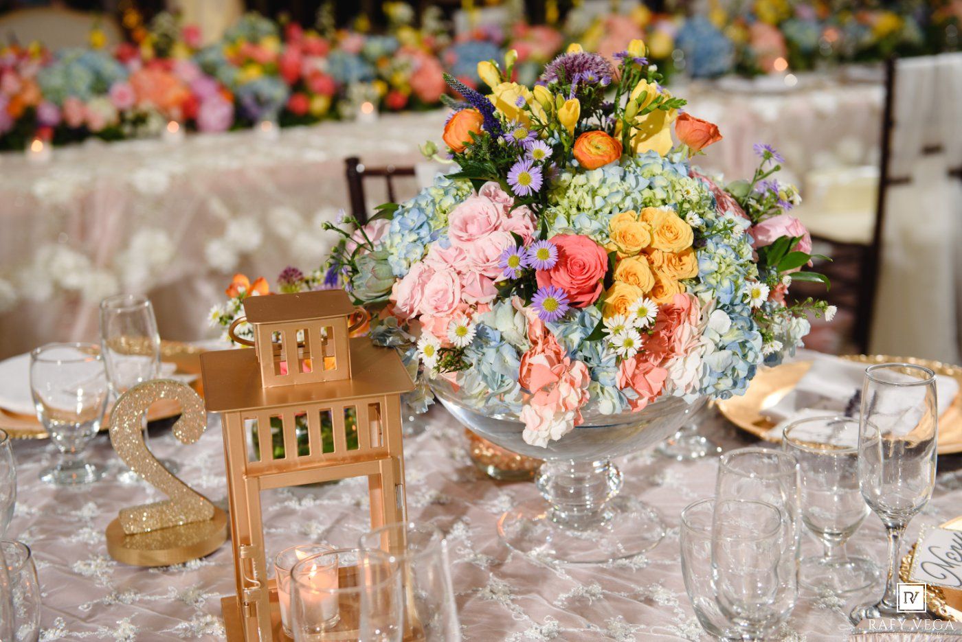 Colorful pastel floral centerpiece with roses and hydrangeas on a wedding reception table with gold accents.