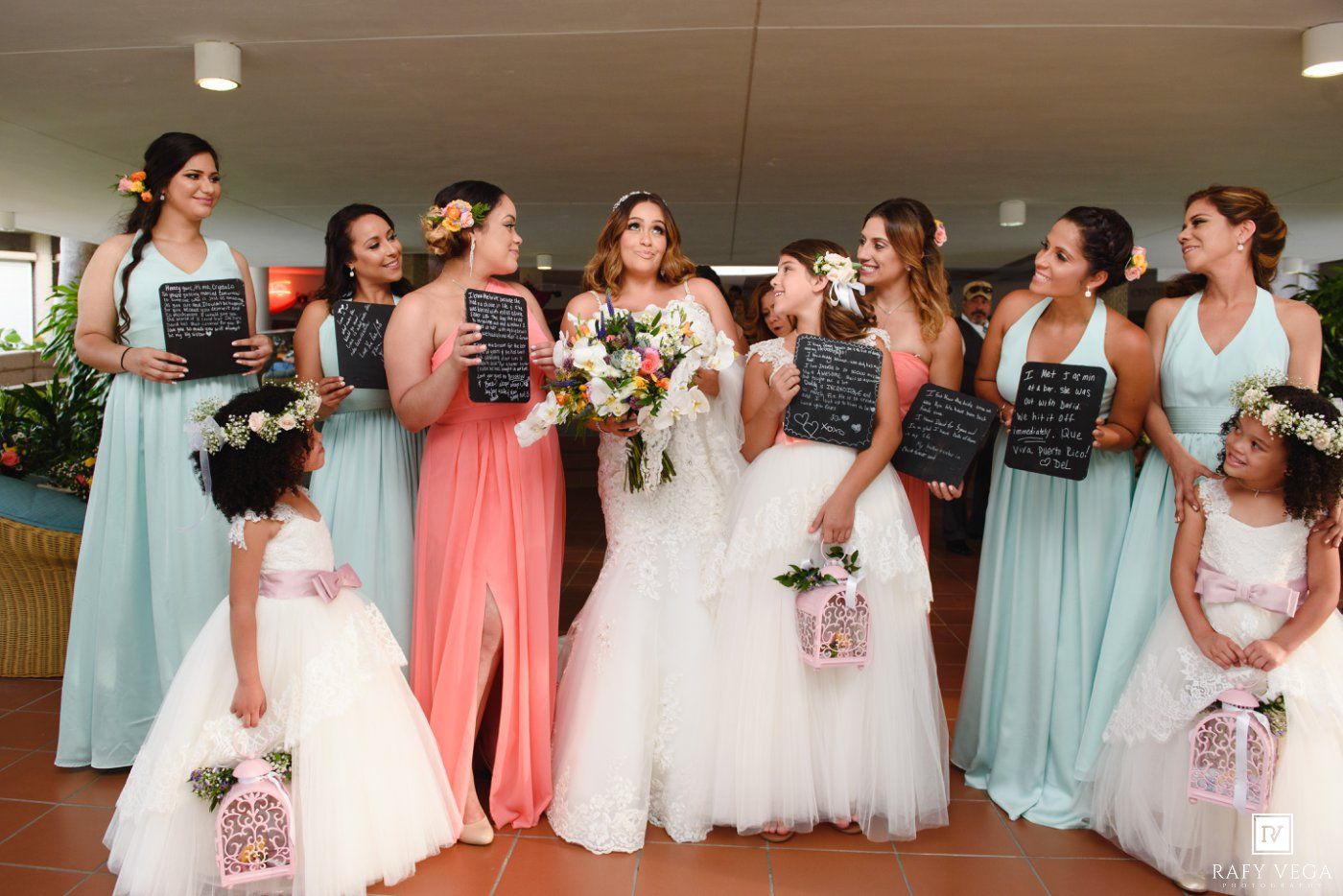 Bride poses with bridesmaids in mint and coral gowns and flower girls in white dresses, holding chalkboard signs indoors.