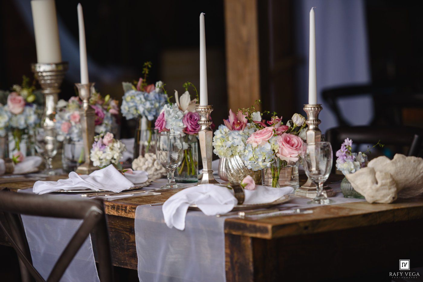 Elegant wedding table setting with white candles, silver candlesticks, and pastel floral arrangements on a rustic wooden table.