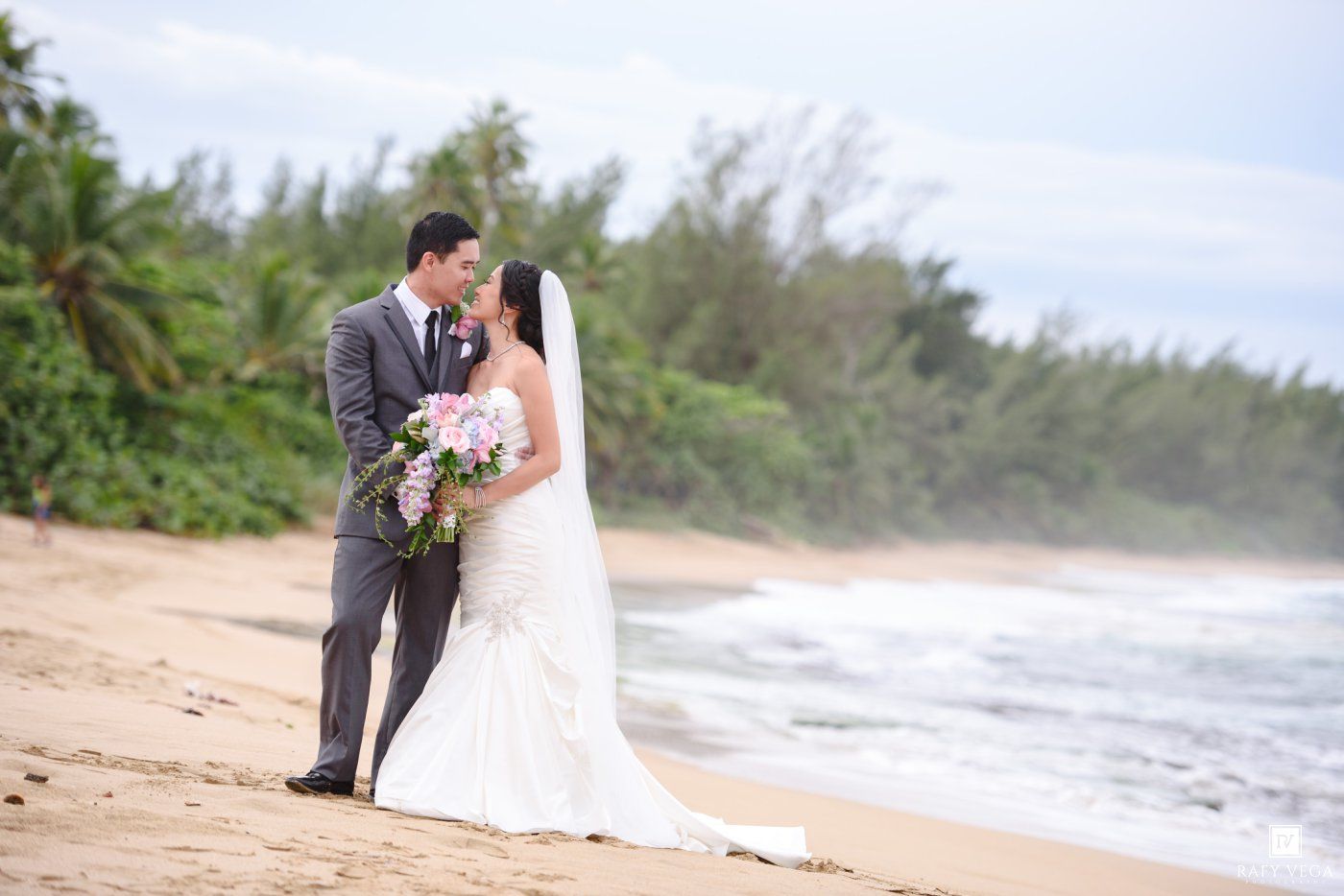Bride and groom share a romantic moment on a tropical beach, with the bride holding a pink bouquet in her wedding dress.
