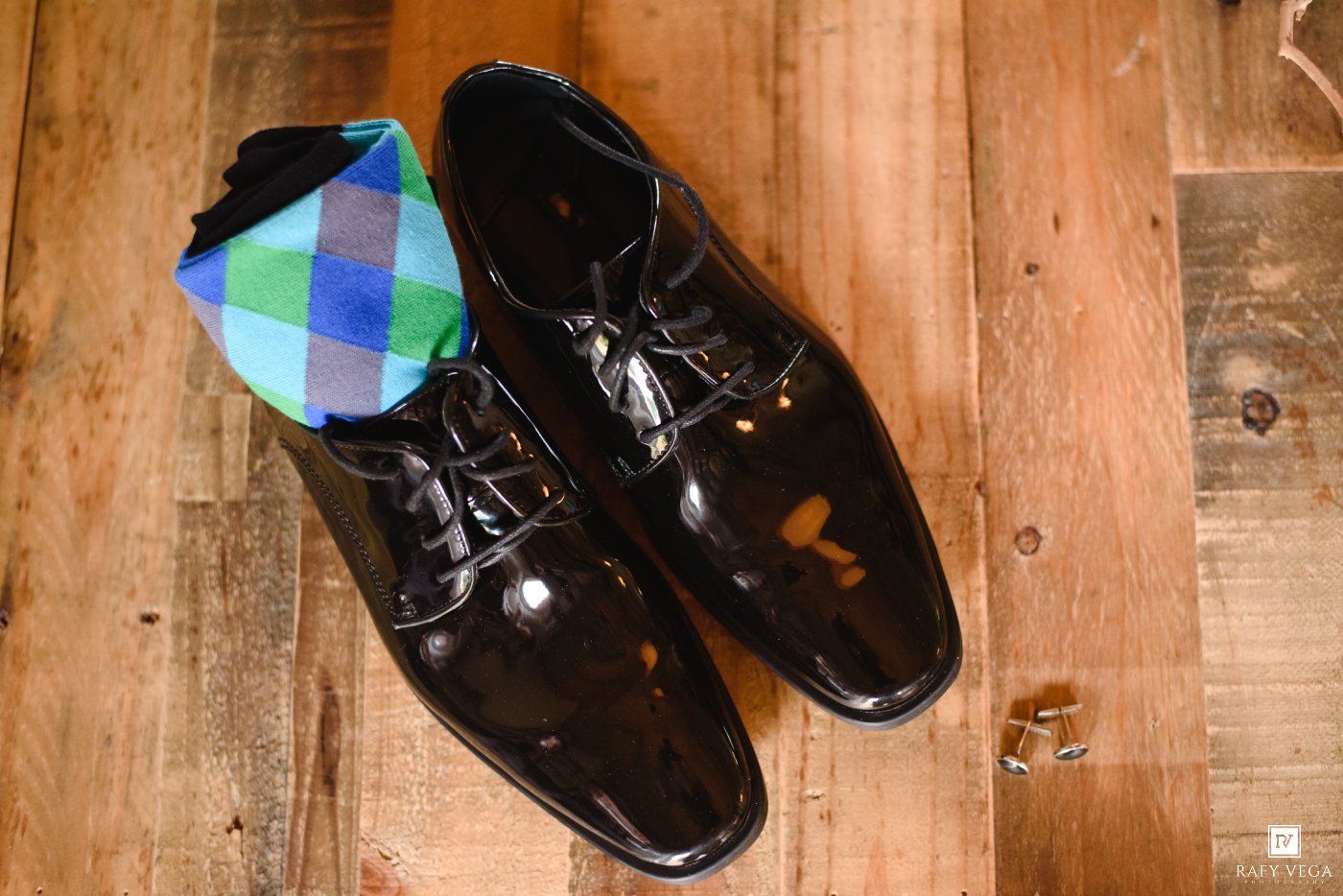 Black patent leather dress shoes with colorful plaid socks on a rustic wooden floor.