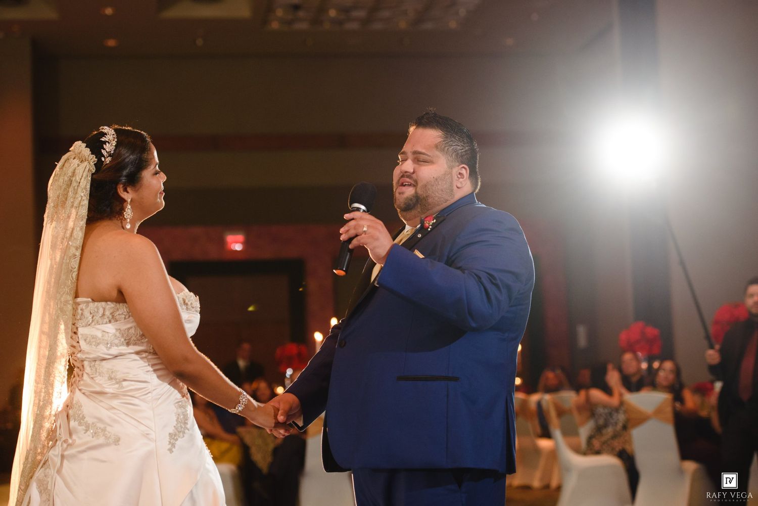Groom in blue suit singing to bride in white wedding gown during romantic first dance at elegant ballroom reception.