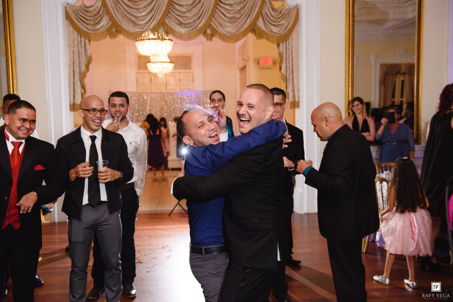 Men laughing and embracing joyfully on the dance floor during a lively wedding reception celebration.