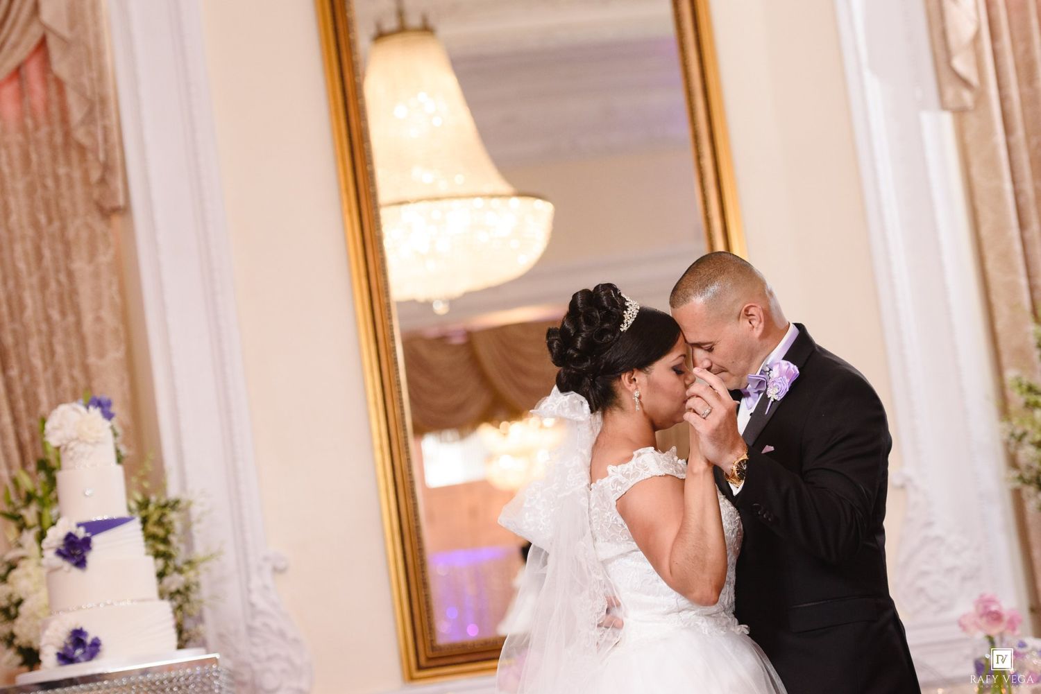 Bride and groom share a romantic kiss near a white wedding cake in an elegant ballroom with gold mirror decor.