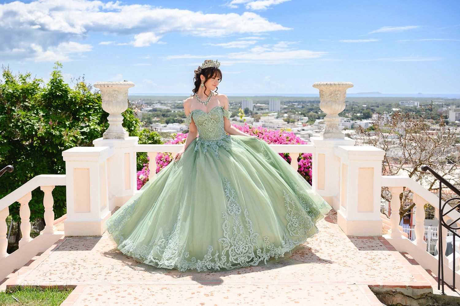 Young woman in a sage green quinceañera ball gown poses on elegant white balcony stairs with city views.