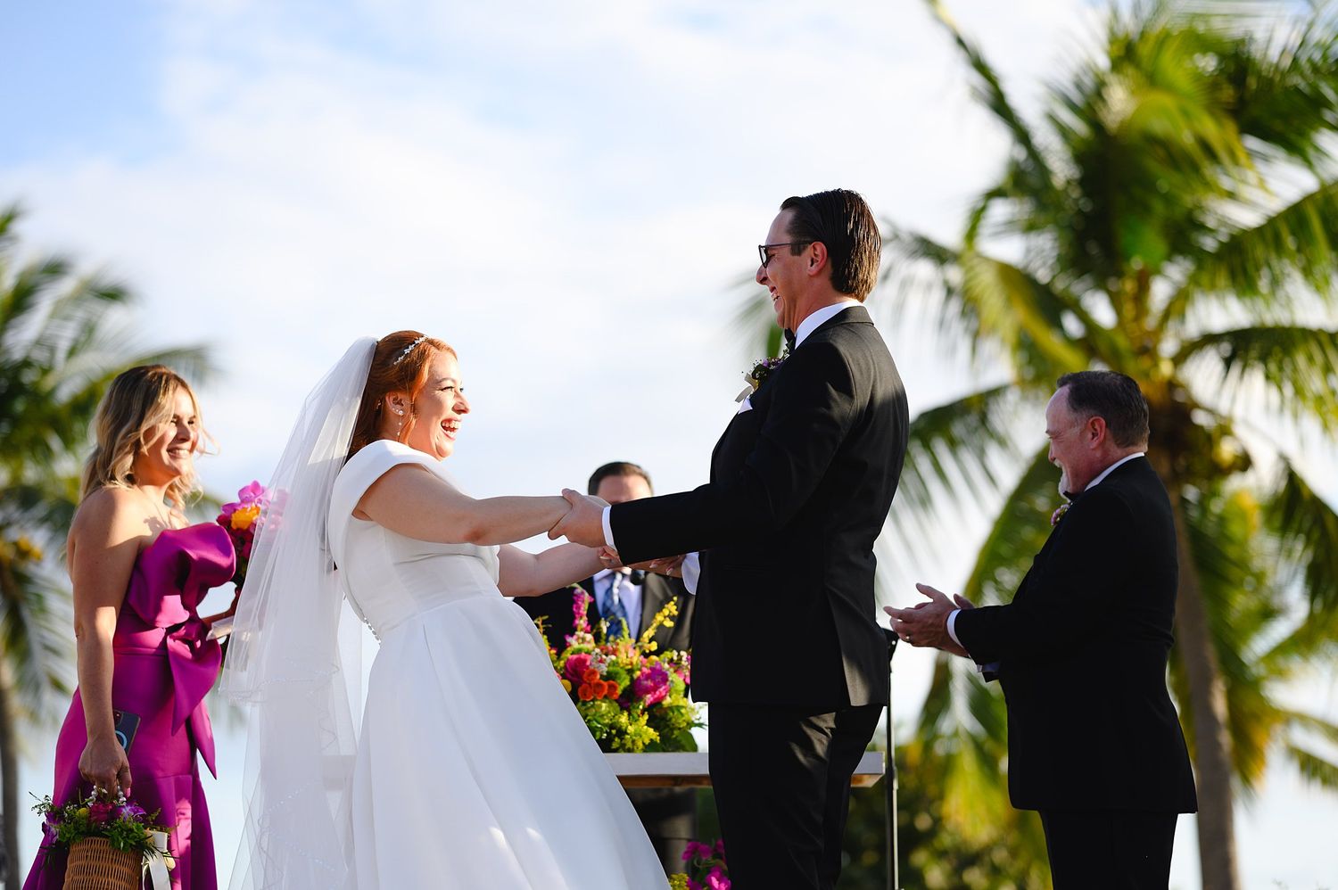 Bride and groom exchange vows at tropical outdoor wedding ceremony with palm trees and colorful flowers.