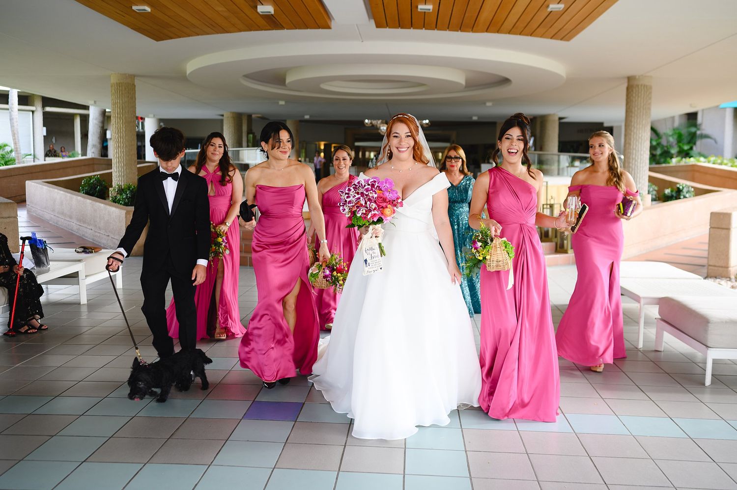 Bride in white gown walks with bridesmaids in pink dresses and groomsman with dog at elegant wedding venue.