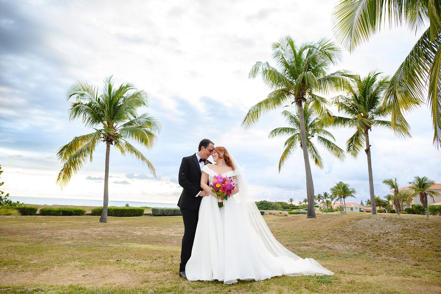 Bride and groom share a romantic kiss surrounded by palm trees near a tropical beach on their wedding day.