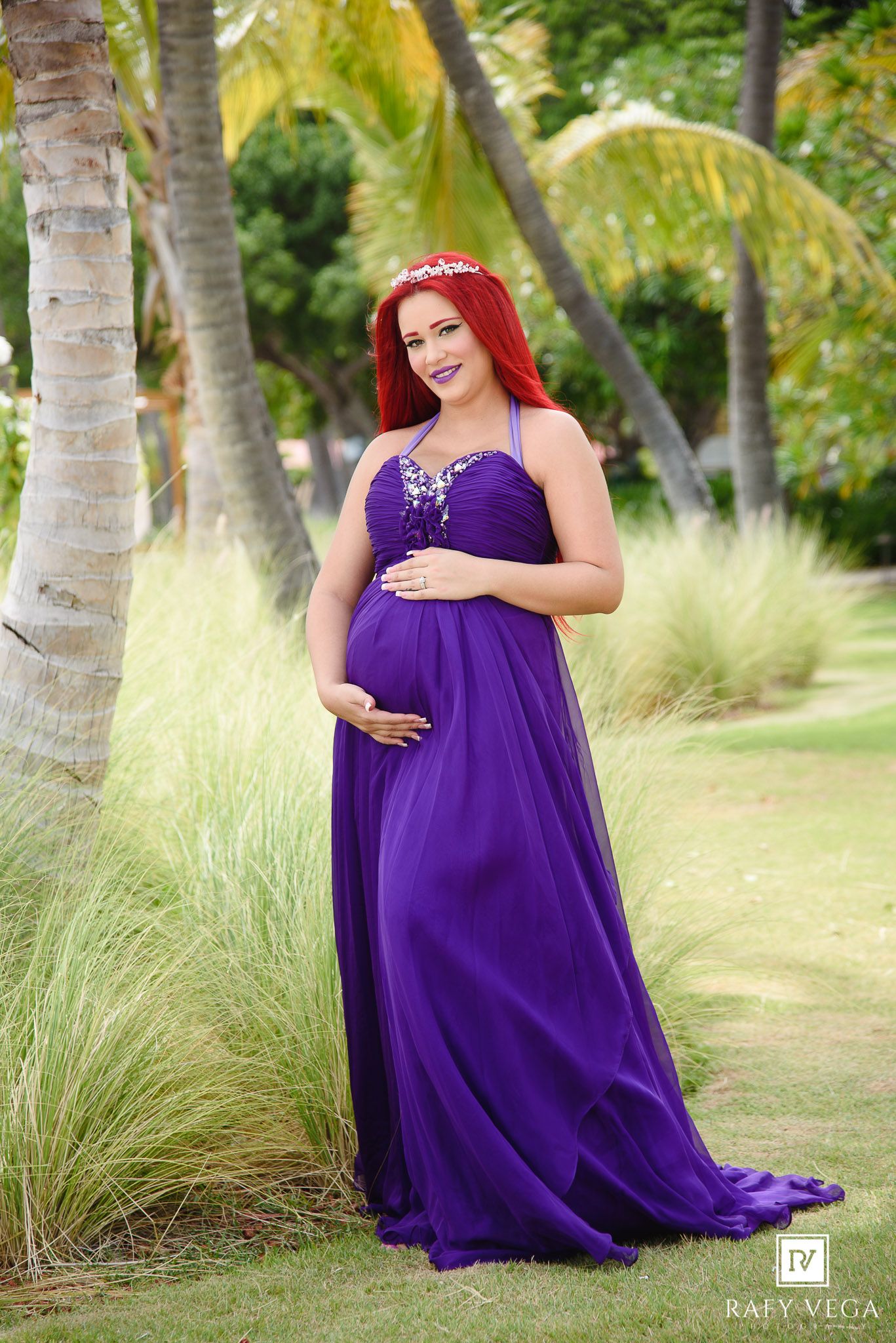 Pregnant woman in purple gown with red hair and floral crown poses gracefully among palm trees in a lush garden.