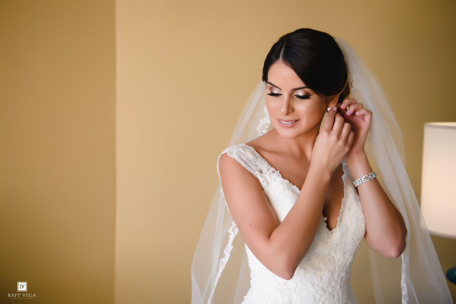 Bride in white lace gown and veil adjusting earring, soft natural light, elegant bridal portrait.