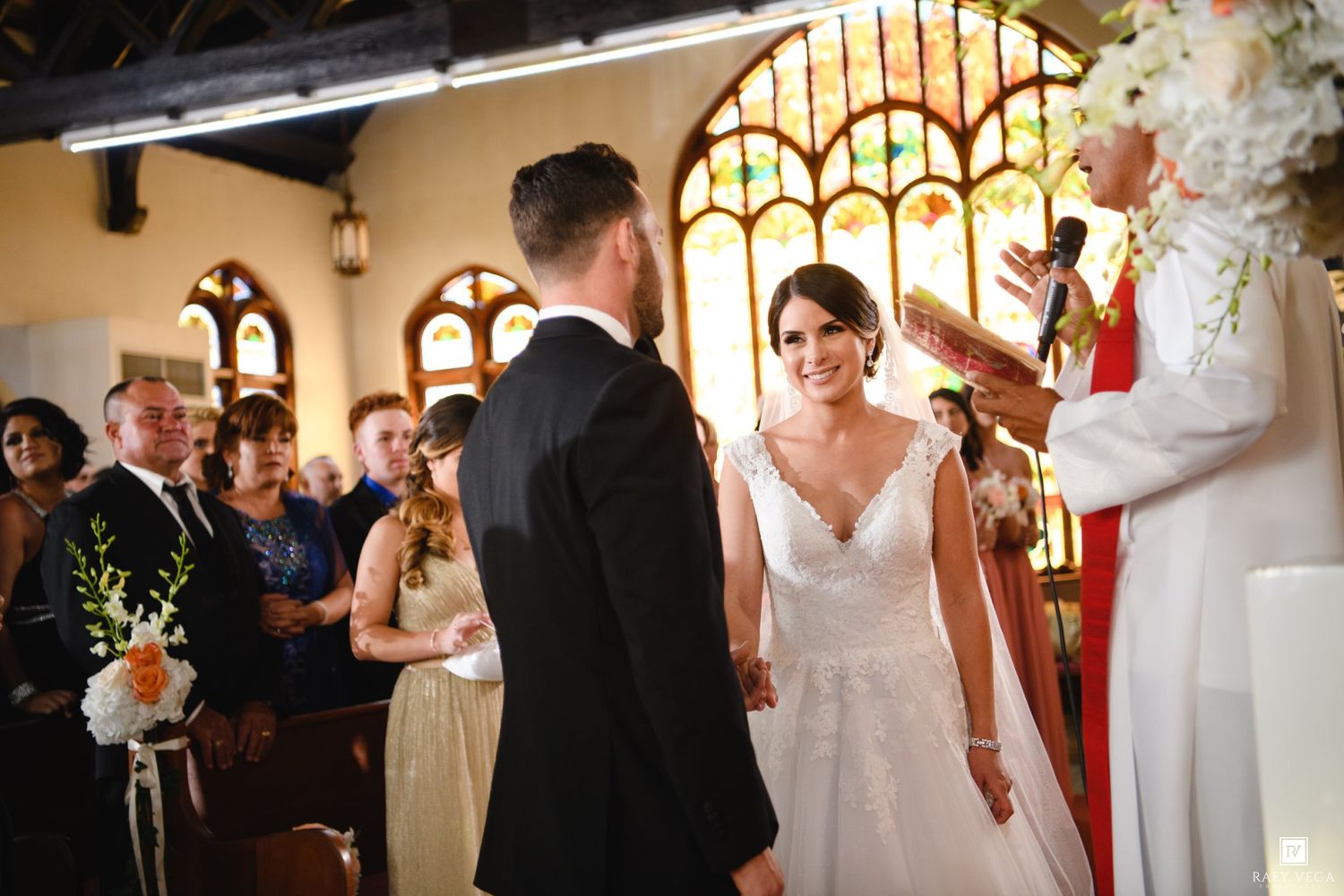 Bride and groom exchange vows at church altar with stained glass windows and officiant during wedding ceremony.