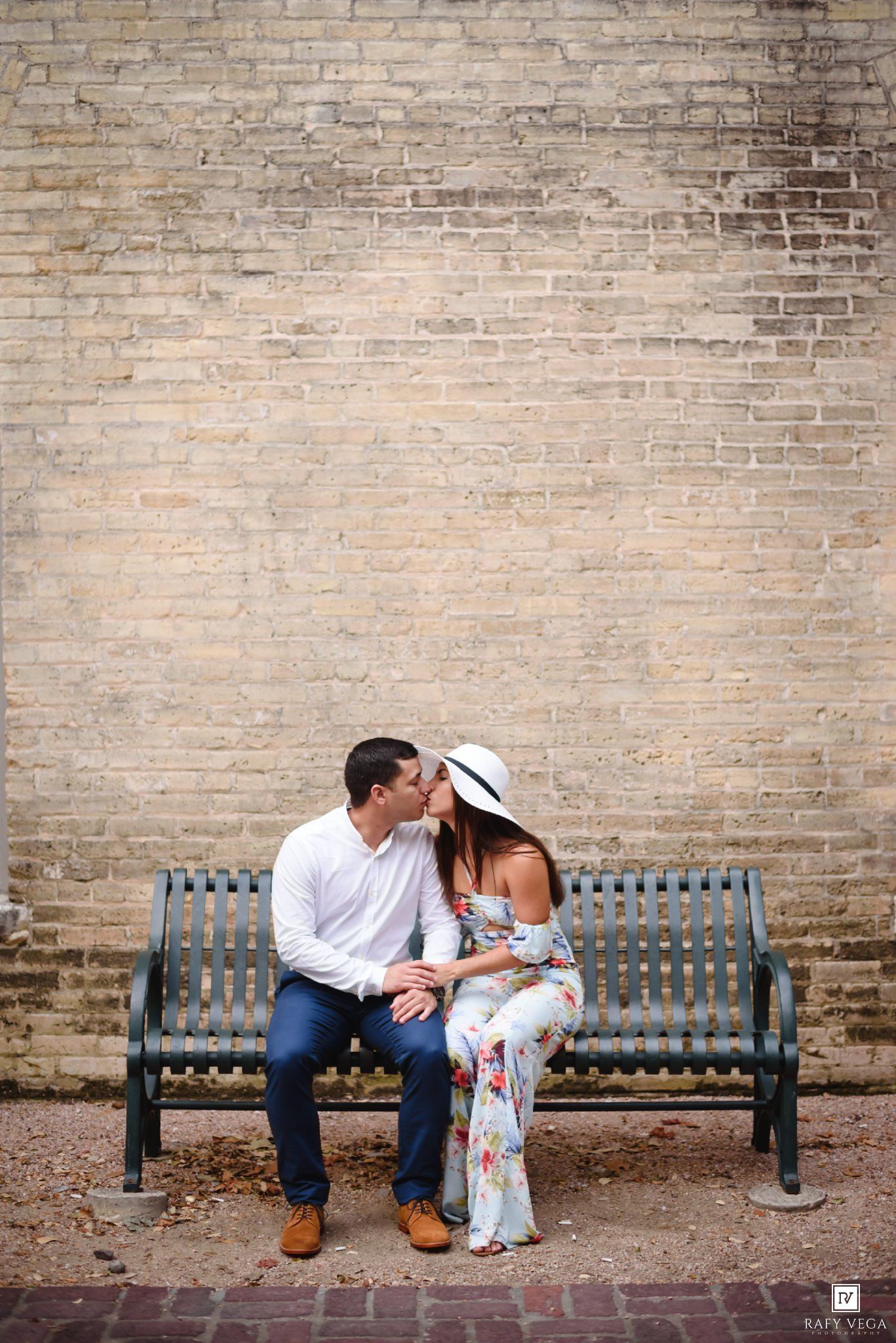 Couple sharing a kiss on a bench against a rustic brick wall, woman in floral dress and white hat.