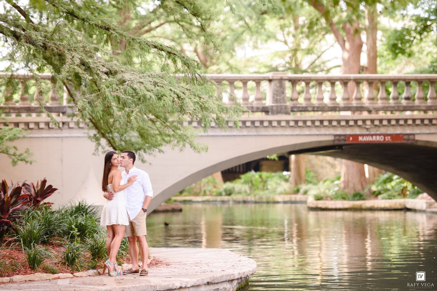 Couple sharing a romantic kiss by a scenic stone bridge over a tranquil river surrounded by lush green trees.