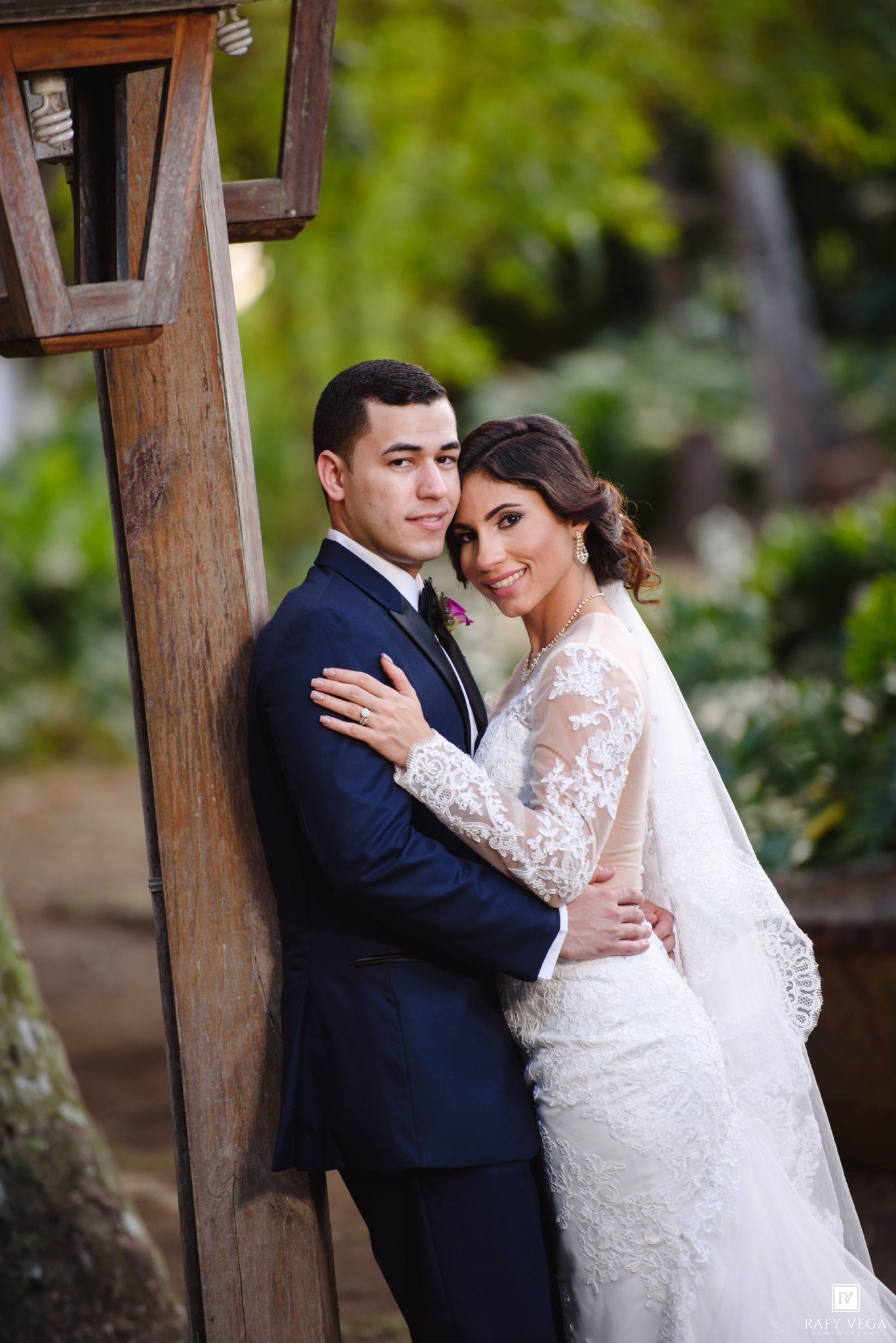 Bride and groom pose elegantly by a wooden post in a lush garden, she in lace gown, he in navy suit.