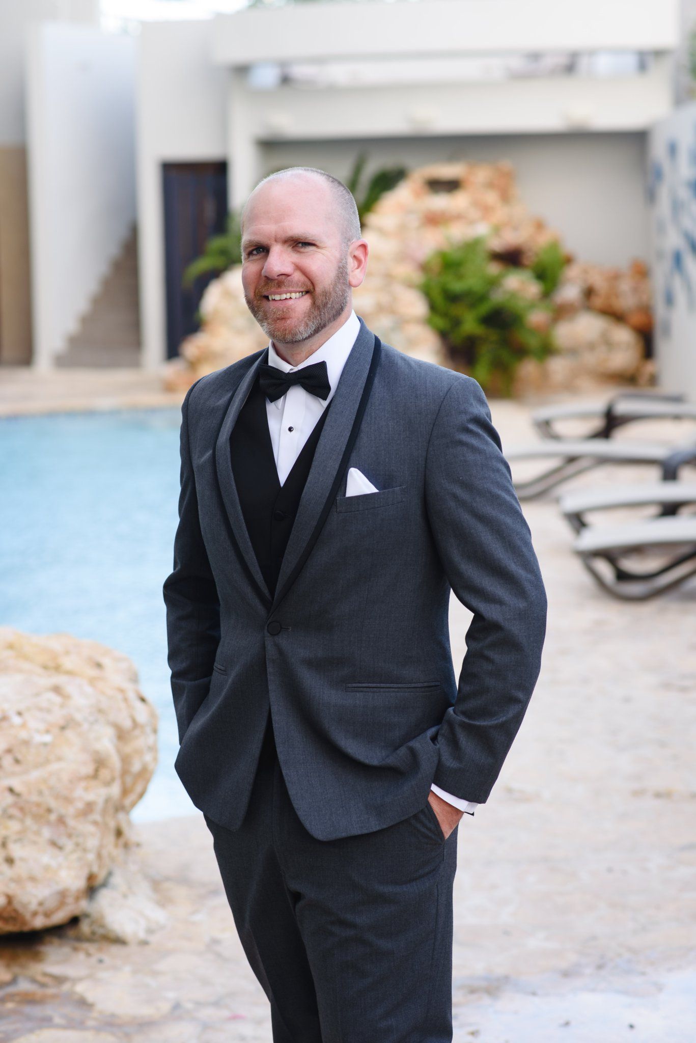 Smiling bald groom in gray tuxedo with black bow tie and white pocket square standing poolside at outdoor venue.
