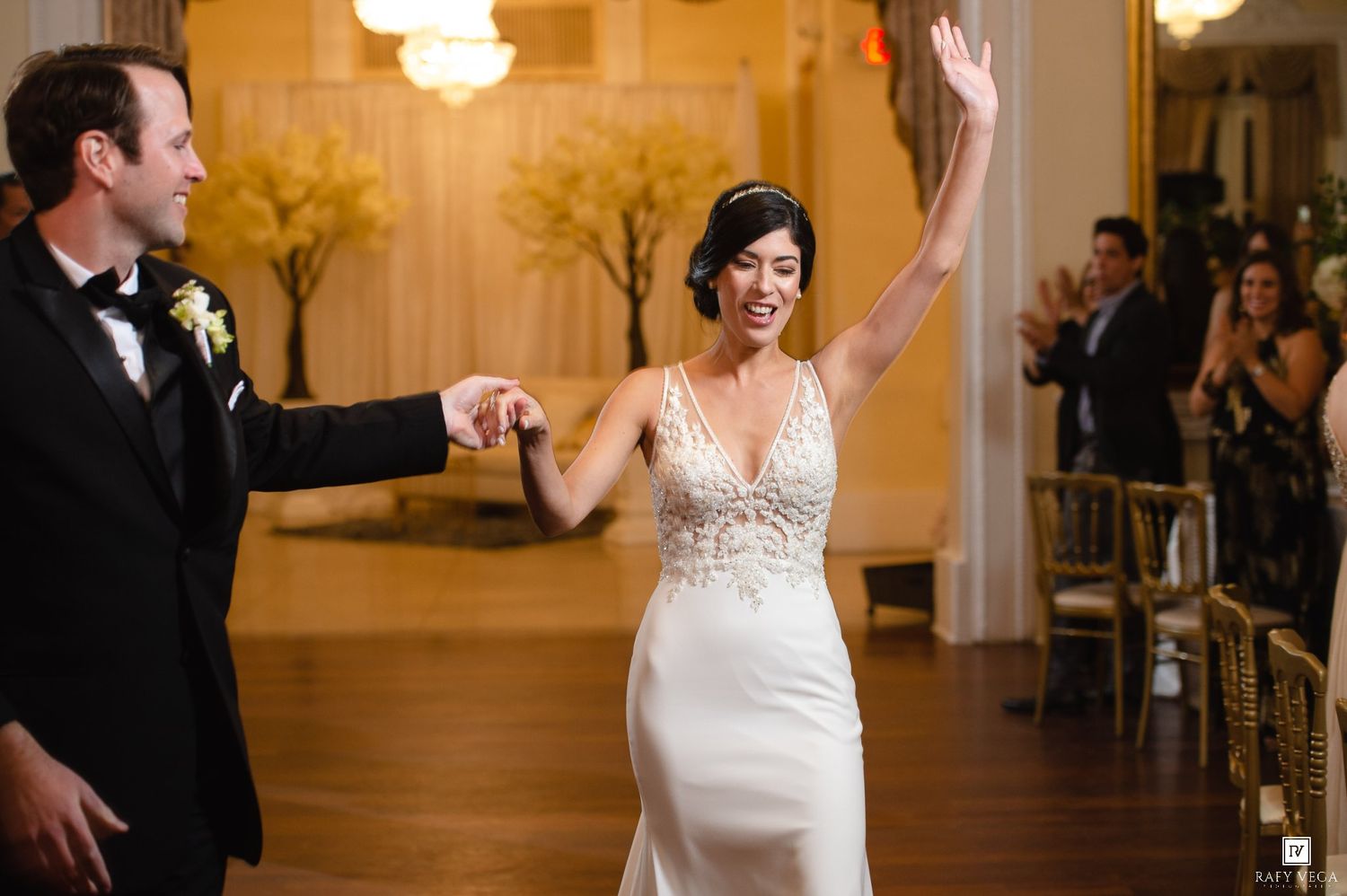 Bride in white gown raises hand joyfully as groom in tuxedo leads her into elegant wedding reception hall.