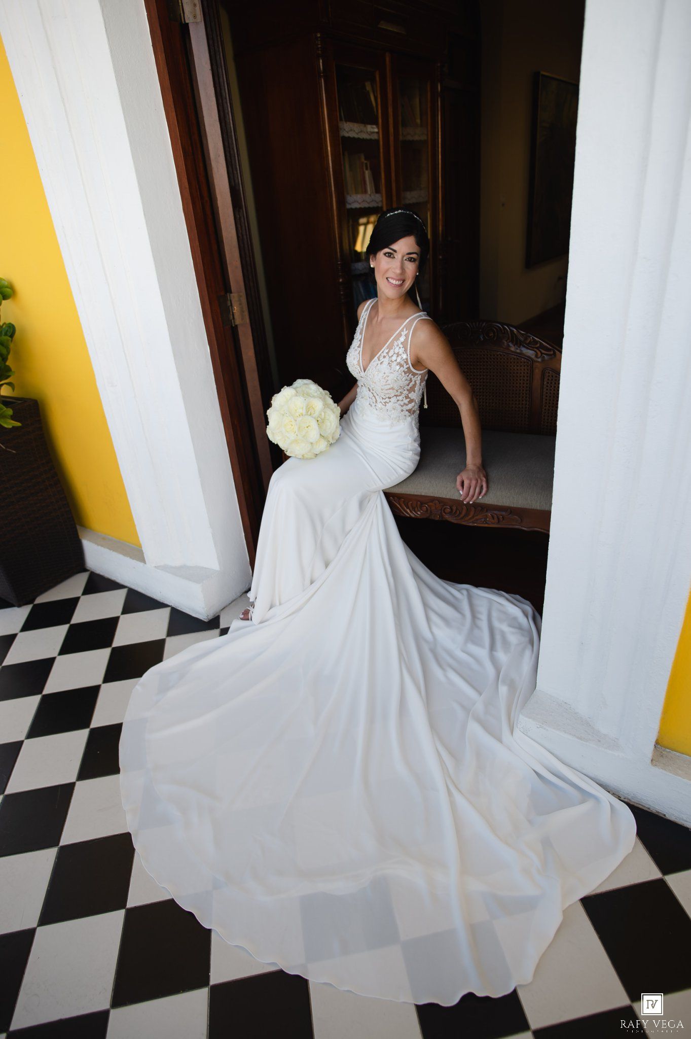Bride in elegant white mermaid wedding gown with lace bodice holding bouquet on black and white checkered floor.