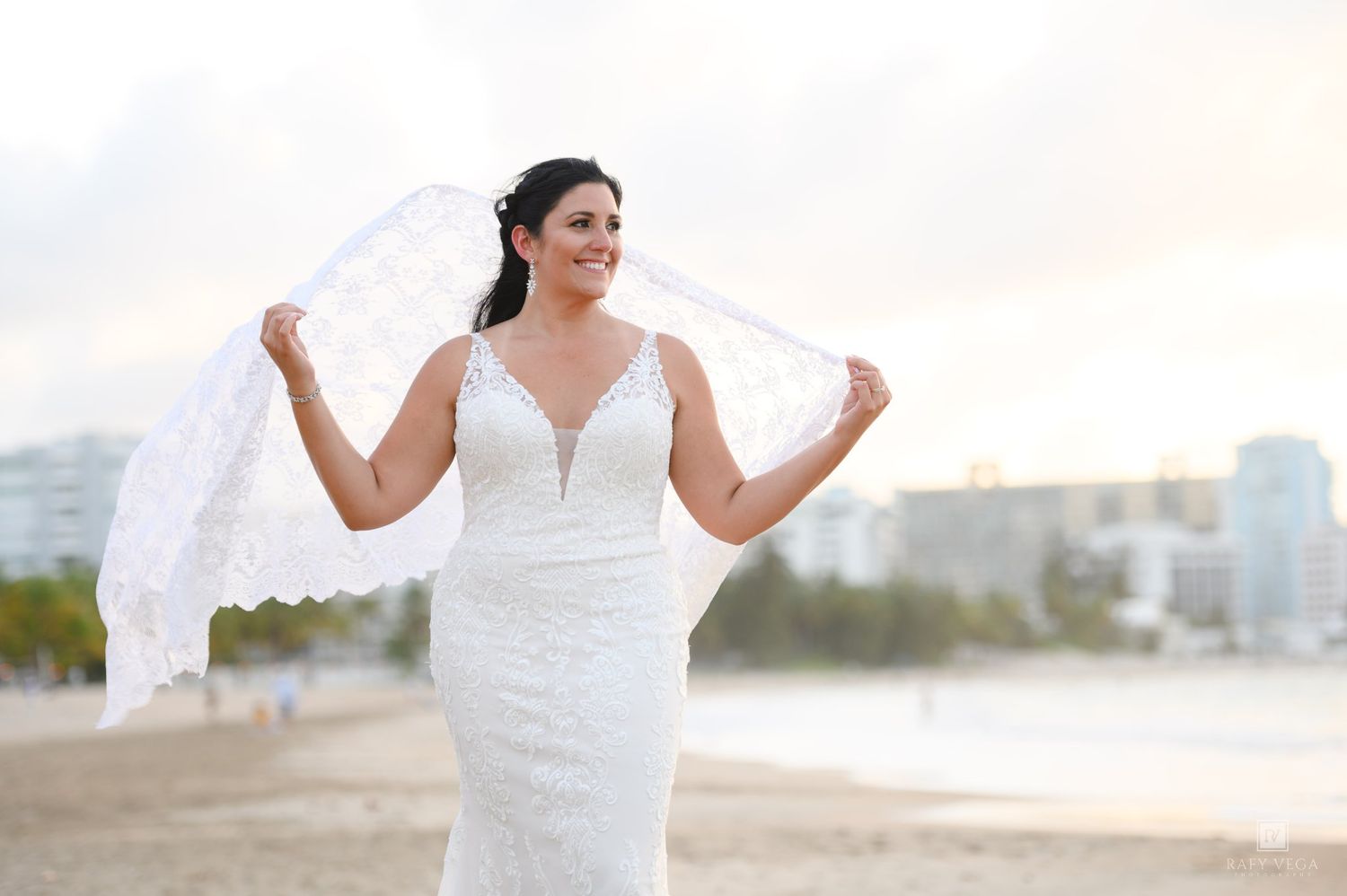 Bride in white lace wedding gown holding veil on beach with city skyline backdrop.