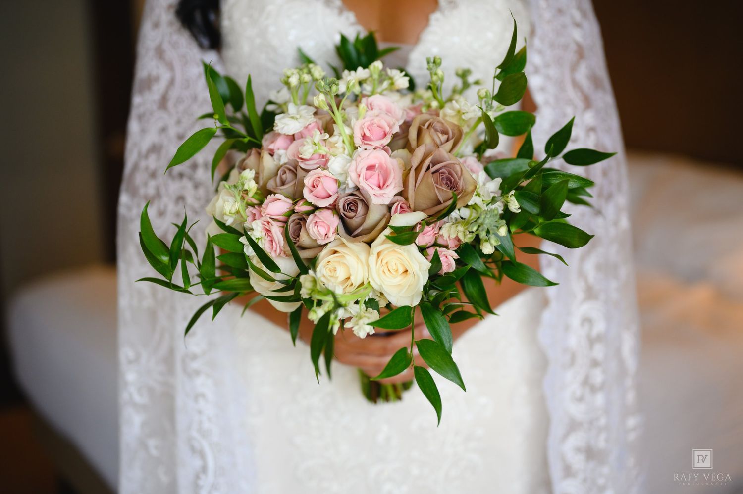 Bride in lace wedding dress holding elegant bouquet of pink roses, cream flowers, and green foliage.